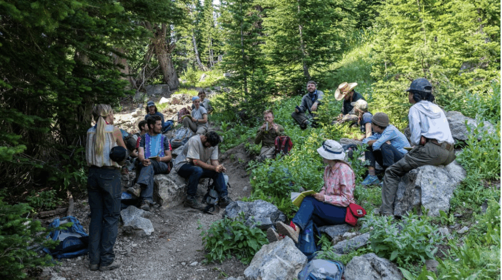 A group of students gathered in the woods and surrounded by trees. Some are sitting on rocks and some in the brush along a dirt path. 